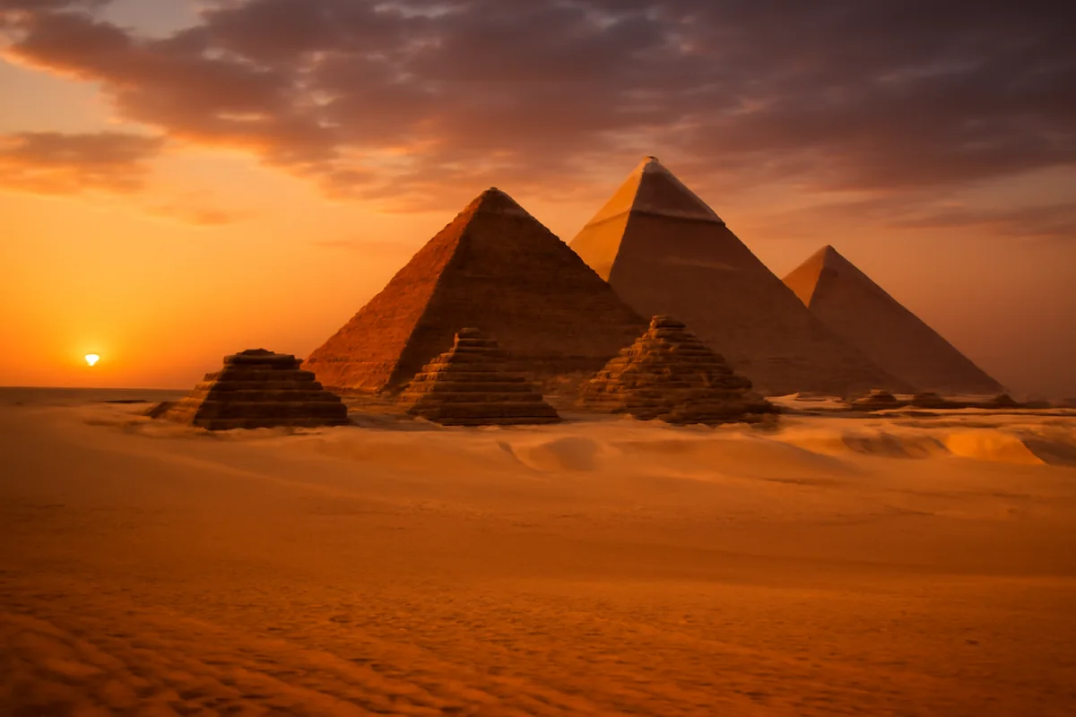 The pyramids of Giza silhouetted against a warm golden sunrise sky, surrounded by sweeping desert sands in Egypt